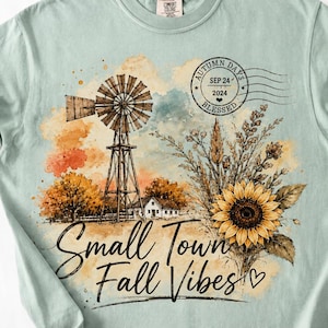 May include: A light sage green t-shirt featuring a watercolor-style graphic of a windmill, a farmhouse, and a sunflower bouquet. The design includes a circular postmark stamp reading AUTUMN DAYS SEP 24 2024 BLESSED and the text Small Town Fall Vibes.