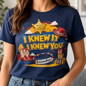 Pode incluir: Uma t-shirt azul-marinho com uma estampa gr&aacute;fica contendo o texto "I KNEW IT I KNEW YOU" e "YOU'VE GOT A FRIEND IN ME". O design inclui uma estrela de xerife dourada, um chap&eacute;u de cowboy vermelho, botas vermelhas e brancas, uma pulseira de contas coloridas e blocos de brinquedo.