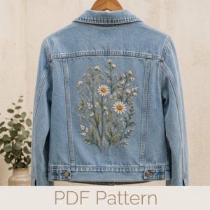 May include: A light blue denim jacket featuring a detailed embroidery design on the back. The embroidery depicts a cluster of wildflowers, including white daisies with yellow centers, green stems, and various small buds and leaves in soft tones.