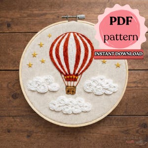 Könnte beinhalten: Gesticktes Heißluftballon-Design in einem Holzreifen. Der Ballon ist rot-weiß gestreift und hat einen geflochtenen Korb. Weiße Wolken und goldene Sterne umgeben den Ballon. Der Text auf dem Bild lautet "PDF pattern" und "Instant Download".