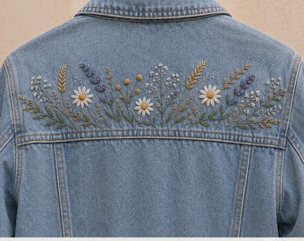 Op de afbeelding: Een lichtblauw spijkerjack met een borduursel van wilde bloemen op het rugpand. Het ontwerp bevat witte madeliefjes, paarse lavendeltakjes, gele knoppen en groen blad. De tekst PDF Pattern is onderaan in de afbeelding zichtbaar.