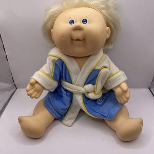 Puede incluir: Una muñeca Cabbage Patch Kid vintage con cabello rubio y ojos azules. La muñeca lleva una bata azul y blanca con ribete amarillo. La muñeca está sentada con las piernas extendidas y los brazos abiertos.