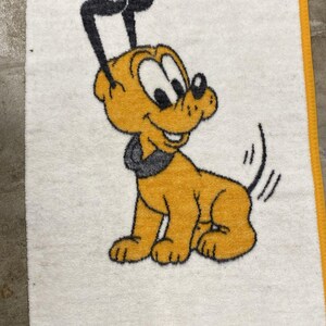 Puede incluir: Una toalla rectangular blanca con un borde amarillo y un gráfico central de un perro de dibujos animados con pelaje amarillo, collar negro y orejas negras. El perro aparece sentado con la cola levantada, representado en un estilo texturizado.