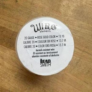 May include: A spool of 20 gauge rose gold color tarnish-resistant wire by The Bead Smith. The white label features the text Wire elements, 20 GAUGE, ROSE GOLD COLOR, 15 YD, and the brand name THE BEAD SMITH printed in black ink on the circular surface.