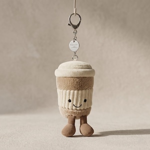 Könnte beinhalten: Ein Schlüsselanhänger in Form einer Plüsch-Kaffeetasse mit einem lächelnden Gesicht und Cordbeinen. Die Tasse ist beige und cremefarben, mit einer weißen gerippten Hülle. Ein kleines silbernes Etikett lautet "have my coffee".