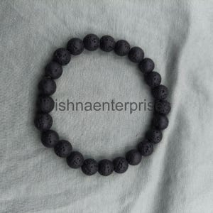 May include: A circular bracelet composed of numerous black, porous lava stone beads strung together. The beads feature a rough, textured surface characteristic of volcanic rock. The text "ishnaenterprise" is visible in the center of the bracelet.