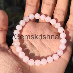 Könnte beinhalten: Ein hellrosa Perlenarmband mit dem Text "Gemskrishna" in der Mitte. Das Armband besteht aus runden, polierten Perlen. Das Armband wird auf der Hand einer Person präsentiert.