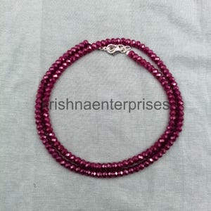 Puede incluir: Un collar compuesto por pequeñas cuentas facetadas de color magenta intenso dispuestas en un patrón circular. La pieza presenta un cierre de mosquetón de metal plateado en la parte superior. El texto "krishnaenterprises" es visible en el centro.