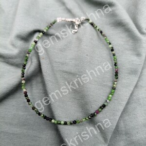 May include: A beaded necklace featuring small, faceted, multi-colored gemstone beads in shades of green, black, and deep red. The necklace is secured with a silver-toned metal lobster claw clasp and a short extender chain at the top center.