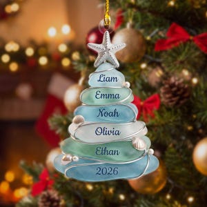 May include: A Christmas tree ornament crafted from stacked, smooth sea glass pieces in shades of blue and green, wrapped in silver wire. The ornament features a silver starfish topper, small white shell accents, and the names Liam, Emma, Noah, Olivia, Ethan, and the year 2026 printed on each layer.