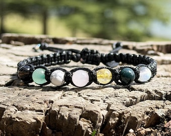 Pode incluir: Uma pulseira de cordão preto com uma fileira de sete contas redondas e lisas de várias cores, incluindo verde-claro, branco, rosa-pálido, amarelo, verde-escuro e branco translúcido, com um fecho de nó de macramé preto ajustável.