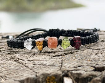 May include: A black macrame bracelet featuring five raw, uncut gemstones in a row. The stones include a clear crystal, a yellow citrine, an orange stone, a green peridot, and a deep red garnet, all secured within the woven black cord band.
