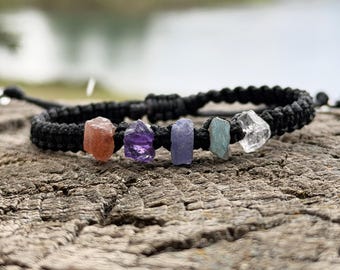 May include: A black woven cord bracelet featuring five small, raw, irregularly shaped gemstones in shades of orange, purple, blue, teal, and clear. The bracelet has a sliding knot closure mechanism and is centered on a textured, weathered wooden surface.