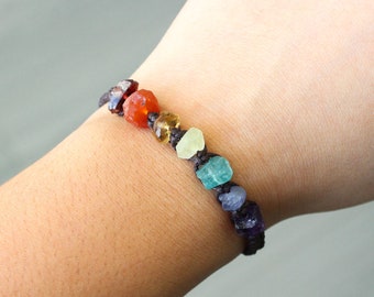Könnte beinhalten: Ein farbenfrohes Edelsteinarmband mit einer Vielzahl von rohen, ungeschliffenen Steinen in Rot-, Orange-, Gelb-, Grün-, Blau- und Lilatönen. Die Steine sind auf einer dunklen Schnur aufgereiht. Das Armband ist ca. 18 cm lang.