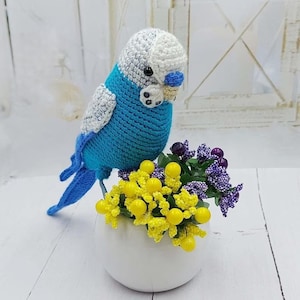 May include: A handmade blue and white crochet parakeet perched on a small white vase filled with yellow and purple flowers. The bird has a blue body, white wings, and a white and gray head. The flowers add a pop of color to the scene.