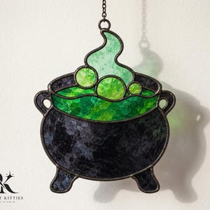 Op de afbeelding: Een glas-in-lood suncatcher in de vorm van een zwarte ketel met drie poten. De ketel bevat borrelende groene vloeistof en opstijgende stoom, gemaakt van getextureerd donker glas en levendige groene glasaccenten, opgehangen aan een metalen ketting.