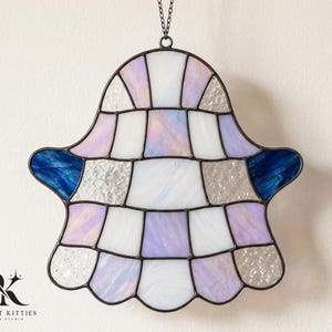 Puede incluir: Un adorno de vitral con forma de mano de Hamsa que presenta una cuadrícula de paneles de vidrio blanco iridiscente, morado claro y transparente texturizado, con detalles de vidrio azul profundo en los dedos laterales y líneas de soldadura oscuras.