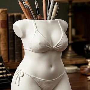 May include: A white ceramic desk organiser shaped like a human torso wearing a bikini. The piece features a textured wave pattern on the bikini top and bottom, with side ties on the bottoms. It is filled with various pens and pencils.