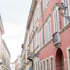 Puede incluir: Una escena callejera con edificios de fachadas rosas y beige. Los edificios tienen numerosas ventanas con contraventanas blancas y balcones de hierro forjado. Las farolas están montadas en los edificios. El cielo está nublado.