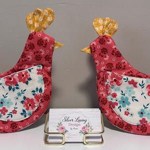Peut inclure: Deux pochettes en tissu en forme de poule avec un motif floral rose sur le corps, une aile blanche à fleurs bleues et roses, et une crête jaune. Un porte-carte de visite est placé entre elles avec une carte indiquant Silver Lining Design by Annie.