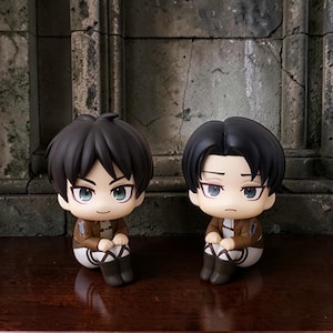 Op de afbeelding: Twee anime-figuurtjes in chibi-stijl met donker haar, grote ogen en een neutrale uitdrukking. Beide figuren dragen bruine jasjes over beige shirts, witte broeken en donkere laarzen, zittend in een houding met de knieën naar de borst getrokken.