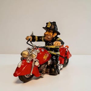 May include: A figurine of a firefighter with a beard wearing a black helmet with FD text, riding a bright red motorcycle. The firefighter wears a black jacket with yellow reflective stripes. The motorcycle features silver accents and a gold emblem.