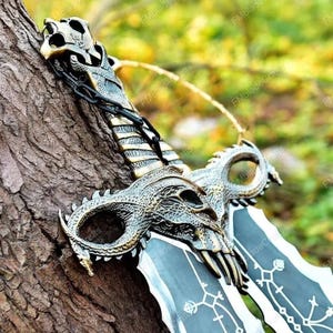 May include: A close-up of a fantasy sword with a dragon-themed hilt and crossguard. The blade is black with white etched designs. The hilt and crossguard are bronze-coloured, featuring intricate dragon skull and scale details. A chain connects the hilt to the pommel.