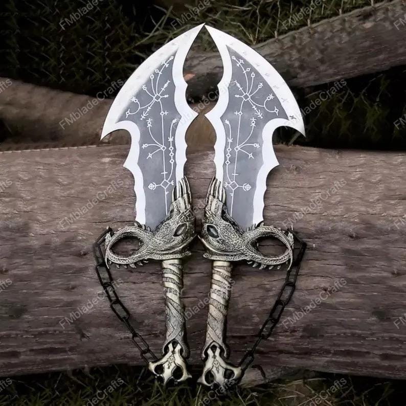 May include: Two silver fantasy swords with dragon-head hilts and curved blades. The blades feature a white design. The swords are linked by a chain and are displayed on a wooden surface.