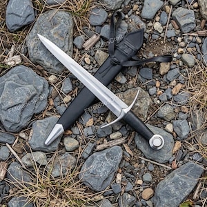 May include: A medieval-style sword with a polished steel blade and a black leather-wrapped handle. The sword rests in a black leather sheath, with a silver tip. The image is set against a backdrop of rocks and dry grass.