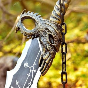 May include: A fantasy sword with a bronze dragon head hilt. The blade is black and white with a geometric design. A black chain is attached to the hilt. The background is blurred. The sword is likely made of metal.