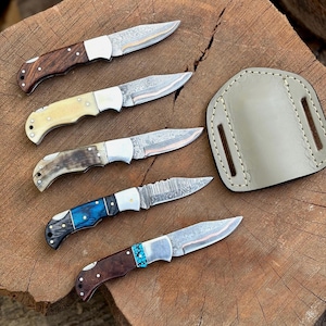 Puede incluir: Una variedad de cuchillos plegables artesanales con diversos materiales de mango, incluyendo madera, hueso y detalles turquesa. Las hojas presentan patrones intrincados y se exhiben sobre una superficie de madera, junto a una funda de cuero.