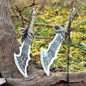 May include: Two decorative swords with detailed handles and blades. The blades are grey with white accents and etched patterns. The handles are gold and silver with a chain. The swords are displayed outdoors, resting against a tree trunk.
