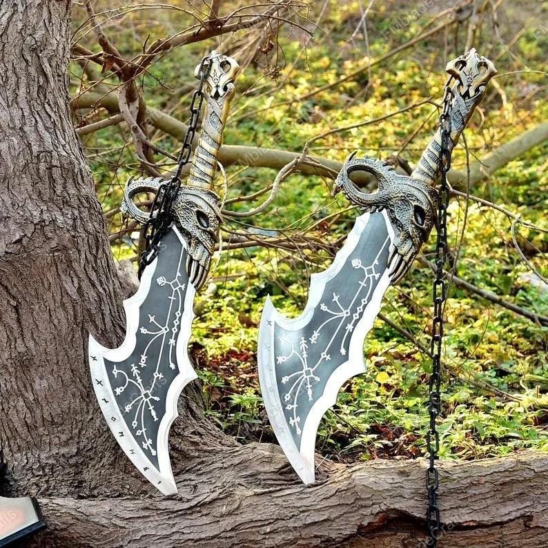 May include: Two decorative swords with detailed handles and blades. The blades are grey with white accents and etched patterns. The handles are gold and silver with a chain. The swords are displayed outdoors, resting against a tree trunk.