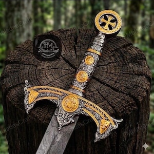 May include: An ornate sword with a silver and gold handle and crossguard. The handle features circular gold accents and a cross design at the pommel. The blade has a patterned design. The sword is displayed on a wooden stump.