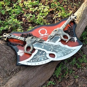 May include: Two silver and black axes with dragon head detailing and a red and black shield featuring "GOD OF WAR" text. The axes are linked by a chain and are displayed on a wooden surface.