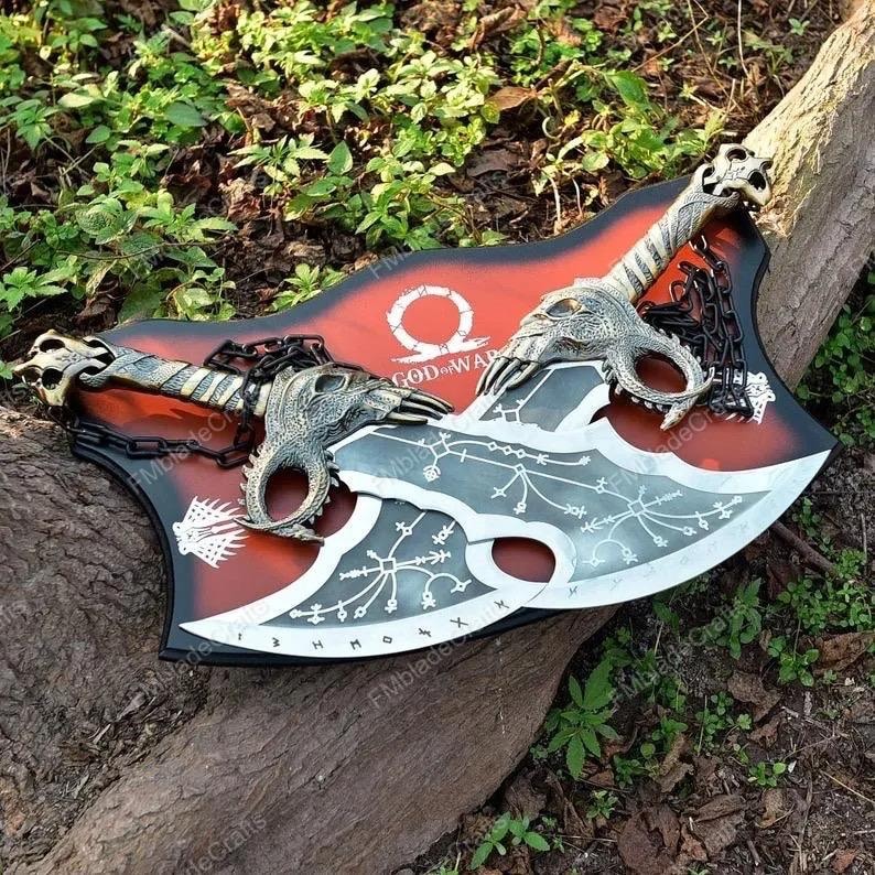 May include: Two silver and black axes with dragon head detailing and a red and black shield featuring "GOD OF WAR" text. The axes are linked by a chain and are displayed on a wooden surface.