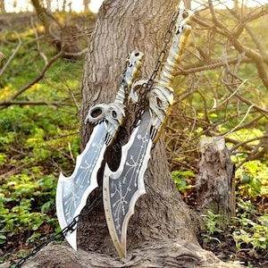 May include: Two silver and gold fantasy blades with skull-shaped hilts and intricate designs. The blades are connected by a chain and are leaning against a tree trunk, set against a backdrop of greenery and sunlight.