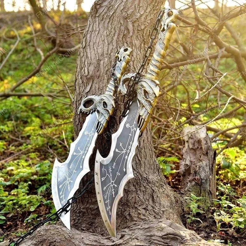 May include: Two silver and gold fantasy blades with skull-shaped hilts and intricate designs. The blades are connected by a chain and are leaning against a tree trunk, set against a backdrop of greenery and sunlight.