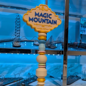 May include: A decorative signpost for Magic Mountain, featuring a cream-colored base and a series of colorful rings in gold, pink, and blue. The sign itself is a light yellow with the words "Magic Mountain" in blue.