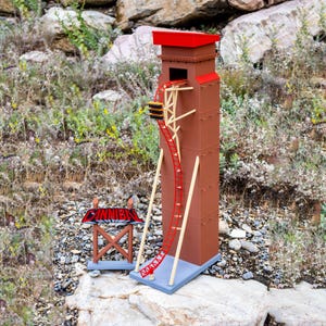 May include: A miniature brown roller coaster tower with a red roof and a red track. A small sign with the word "CANNIBAL" is next to the tower. The model is set on a rocky surface with greenery.