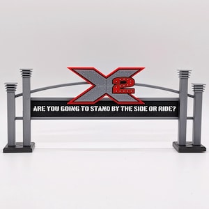 May include: A gray and black 3D printed sign featuring a large red and gray X2 logo at the top. Below the logo, a black rectangular panel displays the white text: ARE YOU GOING TO STAND BY THE SIDE OR RIDE? The sign is supported by four gray pillars.