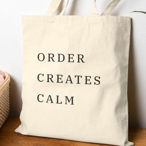May include: A cream-colored canvas tote bag with two shoulder straps, featuring the black text "ORDER CREATES CALM" printed in a simple, sans-serif font on the front center of the bag. The bag is rectangular in shape with a flat bottom.