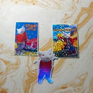 May include: Two rectangular stickers and one die-cut sticker featuring the character Stuart Little. The rectangular stickers display the text Stuart Little and Stuart Little 2 with a glittery finish. The die-cut sticker shows the mouse in a red sweater.