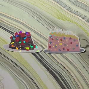 May include: Two stickers of cake slices. One is chocolate with red fruit and colorful accents. The other is pink with star, heart, and glitter details. Both are on gray plates, with a holographic effect.