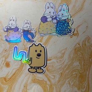 May include: A collection of holographic stickers featuring cartoon characters. The stickers include images of a white rabbit in various outfits and a brown bear-like character holding a rainbow-colored letter. The stickers have a shiny, iridescent finish.