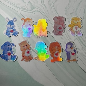 May include: A selection of colourful, holographic stickers depicting cartoon animals. Each sticker has a heart design and is in a different colour, including blue, pink, yellow, and orange. The stickers are on a light green background.