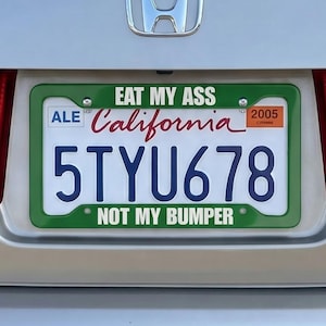 Puede incluir: Parte trasera de un coche plateado con una matrícula de California. La matrícula dice "5TYU678" e incluye el texto "EAT MY ASS" y "NOT MY BUMPER". El coche tiene luces traseras rojas.