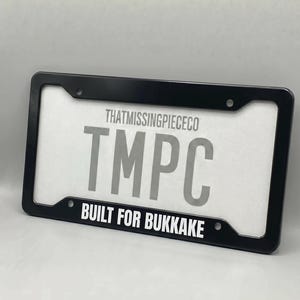 Puede incluir: Marco de matrícula negro con un inserto blanco. El inserto presenta el texto "THATMISSINGPIECECO" encima de las letras "TMPC" y la frase "BUILT FOR BUKKAKE" debajo.
