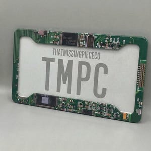 Op de afbeelding: Groen kentekenplaatframe met een printplaatontwerp. Het frame heeft de tekst "THATMISSINGPIECECO" en "TMPC" in grijs. Het frame is rechthoekig en heeft een wit midden.