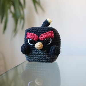 Può includere: Un personaggio di Angry Birds fatto a mano all'uncinetto, nero, con sopracciglia rosse, becco beige e occhi bordati di bianco. Il piccolo peluche rotondo ha una piccola coda con la punta gialla. Il giocattolo è appoggiato su una superficie riflettente.